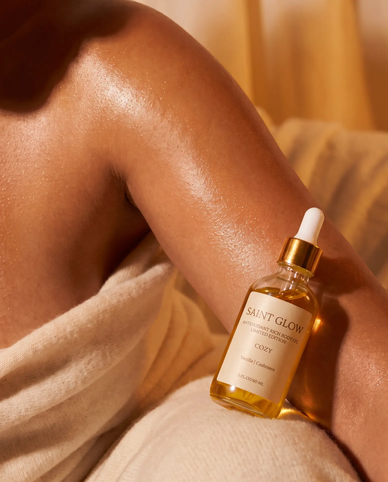 Saint Glow Cozy body oil resting on glowing, oiled skin beside a soft cream knit